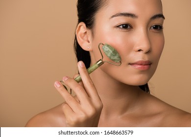beautiful naked asian girl massaging face with jade roller isolated on beige