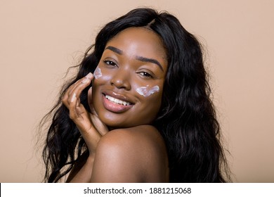 Beautiful Naked Black African Model With Flawless Skin And Long Hair, Applying Moisturiser Face Cream On Her Cheeks, Posing To Camera With Smile Against Beige Background. Beauty, Cosmetics, Skincare