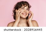 Beautiful natural girl smiling, rubbing her face with facial cleanser for glowing healthy skin, looking happy. Curly young woman showering, using skincare hydrating products, pink background