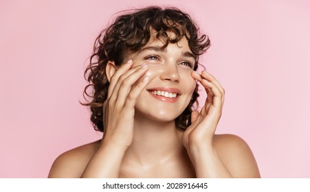 Beautiful natural girl smiling, rubbing her face with facial cleanser for glowing healthy skin, looking happy. Curly young woman showering, using skincare hydrating products, pink background - Shutterstock ID 2028916445