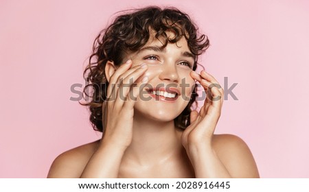 Beautiful natural girl smiling, rubbing her face with facial cleanser for glowing healthy skin, looking happy. Curly young woman showering, using skincare hydrating products, pink background