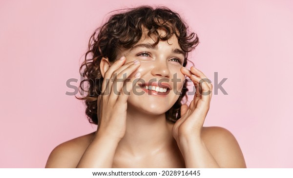 Beautiful natural girl smiling, rubbing her\
face with facial cleanser for glowing healthy skin, looking happy.\
Curly young woman showering, using skincare hydrating products,\
pink background