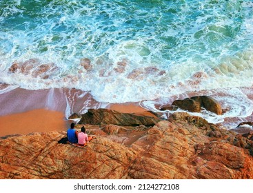 Beautiful nature scene - raging sea, green water, white surf foam and pair of boy and girl sit on rocks. Stormy weather in Costa Brava, Mediterranean sea, Calella beach, Barcelona, Catalonia, Spain