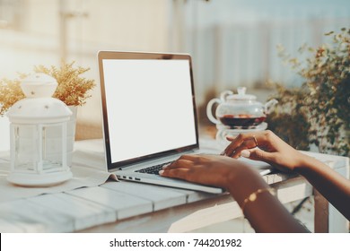 Beautiful and neat hands of African American female with beige manicure, using laptop with white blank screen mock-up in street cafe with glass teapot in background; wooden white table, sunny day