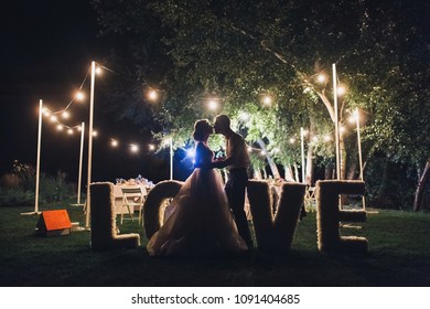 Beautiful Newlyweds Kiss Tenderly At A Wedding Party With Lamps. Stylish Wedding Ceremony. Love In The Frame. Designer Letters. Grain, Film Effect.