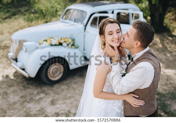 Beautiful newlyweds are smiling against the\
background of an old retro car and summer nature. Wedding portrait\
of a stylish, smiling groom and lovely bride with curly hair.\
Photography and\
concept.