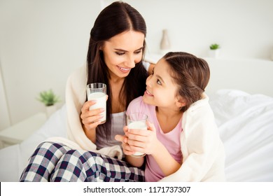 Beautiful nice-looking charming kind mum spending free time with her sweet small little daughter in nightwear, drinking healthy dairy beverage, sitting on bed, covered with blanket in light interior - Powered by Shutterstock