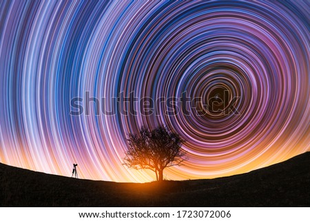 Beautiful night landscape, The camera on the tripod and tree silhouette on the hill under the  colorful star trails on the sky. Night timelapse photography.