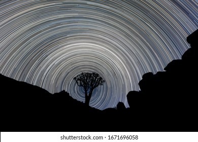 A Beautiful Night Sky Photograph Of A Silhouetted Quiver Tree Framed By Rocky Mountains, With Circular Star Trails Creating A Vortex Around The Tree, In The Richtersveld National Park, South Africa.