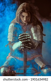 A beautiful noble warrior woman in chain mail and plate armor sits, leaning on her sword and bowing her head. Medieval knight.