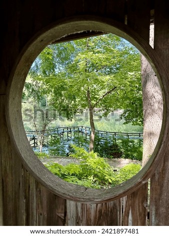 Beautiful oasis of lush green trees, magical bridge and water captured through a circular weathered wood port hole window.