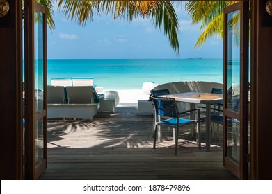 Beautiful ocean view from the window. Turquoise sea, white beach furniture and palm leaves and white sand.