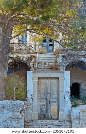 Beautiful old and abandoned mansion, with a traditional wooden front door and staircases leading to the main house building, and a large pine tree in its garden, in Mesaria village, Santorini, Greece.