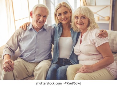 Beautiful Old Couple And Young Girl Are Hugging, Looking At Camera And Smiling While Sitting On Couch At Home