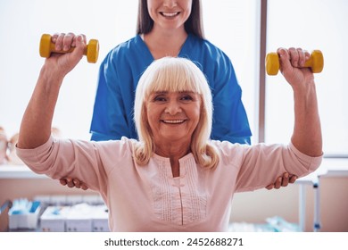 Beautiful old lady is doing exercises with dumbbells, looking at camera and smiling, in hospital ward. Attractive nurse is helping her - Powered by Shutterstock