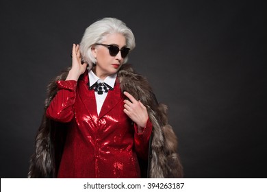 Beautiful old woman with blond hair wearing sunglasses. Fashionable lady in red business suit and expensive fur coat looking like Devil wears Prada.