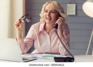 Beautiful Old Woman In Classic Shirt Is Talking On The Phone, Holding Eyeglasses And Smiling While Working With A Laptop At Home