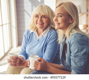 Beautiful Old Woman And Young Girl Are Drinking Tea, Talking And Smiling While Sitting On Couch At Home