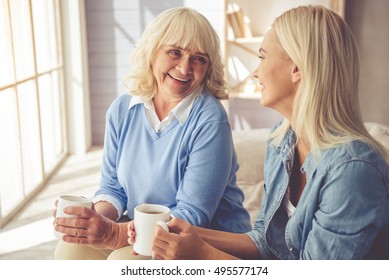 Beautiful Old Woman And Young Girl Are Drinking Tea, Talking And Smiling While Sitting On Couch At Home