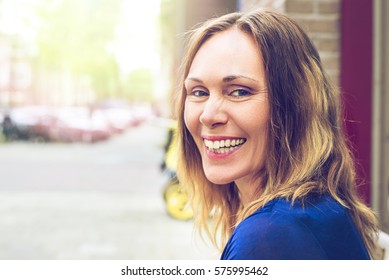 Beautiful Older, Middle Aged Woman Is Happy And Laughing On A Sunny Day In A European City. Wearing A Blue Dress. A Blurred Background, Space For Copy. A Vintage, Yellowish Feel To It.   