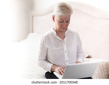 Beautiful Older Woman With A Short Haircut Works Behind A Laptop On A Bed In Her Room