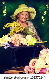 Beautiful Older Woman Sitting In The Garden Among The Flowers.