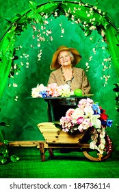 Beautiful Older Woman Sitting In The Garden Among The Flowers.