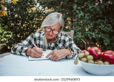 Beautiful older woman is sitting in the garden and writing in her diary. Female senior is keeping daily journal, writing down thoughts and feelings. - Powered by Shutterstock