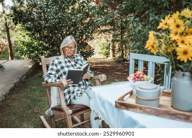 Beautiful older woman is sitting in rocking chair in the garden and reading book. Female senior is enjoying a peaceful moment at home, savoring her freedom. - Powered by Shutterstock