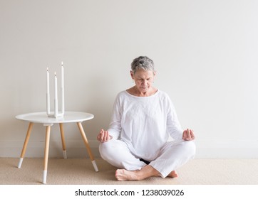 Beautiful Older Woman In White Clothing Sitting In Yoga Meditation Position With Candles Against Neutral Wall Background (selective Focus)