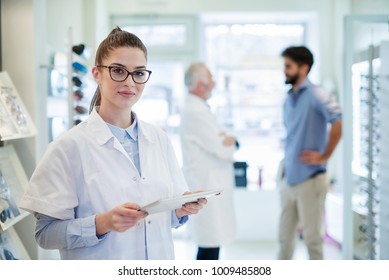 Ein schöner Optiker steht im Geschäft mit Tablet-Computer, während der Arzt im Hintergrund mit dem Patienten spricht.