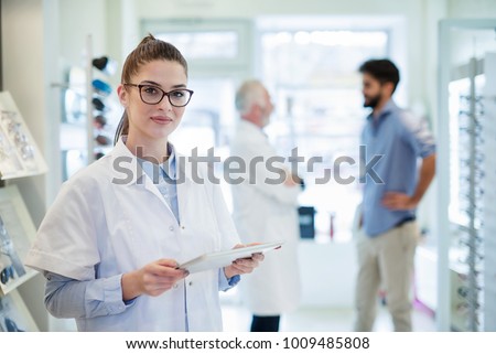 Beautiful optician is standing in the store with tablet computer while the doctor is talking in the background with patient.