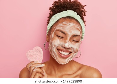 Beautiful optimistic Afro American woman cleanses face with foam, refreshes skin, has well cared complexion, holds heart shaped sponge for beauty procedures, stands bare shoulders with closed eyes - Powered by Shutterstock