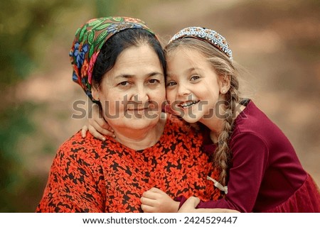 A beautiful oriental grandmother sits on a blanket with her European granddaughter. The granddaughter hugs her grandmother and smiles. Turkmen grandmother with a girl close-up.