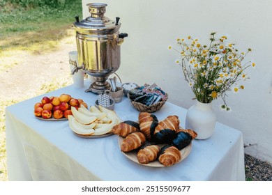 A beautiful outdoor breakfast is arranged with a traditional samovar, croissants and pastries displayed on a table, alongside fresh fruits and a vase of flowers. - Powered by Shutterstock