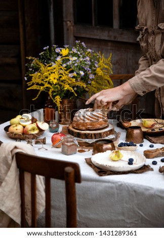 beautiful outdoor still life in country garden with bundt cake on wooden stand on rustic table with tablecloth, copper cups, berries, apples, cinnamon, plates, bouquet, chairs. woman cut the cake