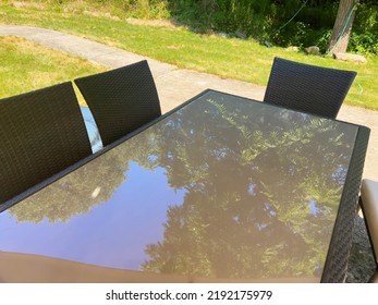 Beautiful Outdoor Summer Table With Glass Top Reflecting The Green Leaves On The Tall Trees Above And Blue Sky In A Green Grass Yard With Cememt Walkway. Three Chairs Are Visible With No People, And