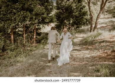 A beautiful outdoor wedding scene where the bride and groom hold hands and walk through a scenic, wooded path. The bride is dressed in a flowing white gown, and the groom is in a light-colored suit.  - Powered by Shutterstock