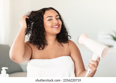 Beautiful oversize woman in bath towel using hairdryer after morning shower and smiling at camera, copy space. Body positive lady drying her hair, making hairdo. Everyday hygiene concept