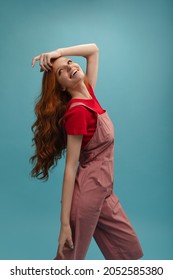 Beautiful pale-skinned girl with radiant smile stands with her head thrown back and looks into distance. She has long red wavy hair and is wearing T-shirt and jumpsuit. Beauty and youth concept