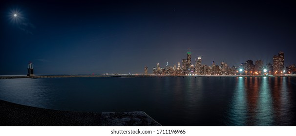 A Beautiful Panoramic Shot Of The Chicago Skyline And City At Night From The Curved Pier At North Avenue Beach With Light Radiating From The Moon And Lights Reflecting On The Water Of Lake Michigan.