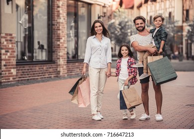 Beautiful Parents And Their Cute Little Kids Are Holding Shopping Bags, Looking At Camera And Smiling While Standing Outdoors