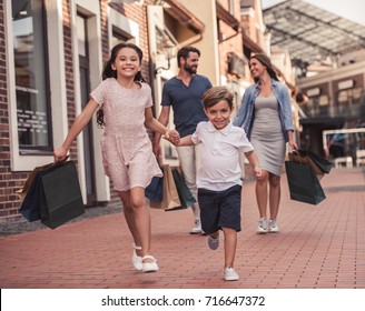 Beautiful Parents And Their Cute Little Kids Are Carrying Shopping Bags, Holding Hands And Smiling While Walking Down The Street