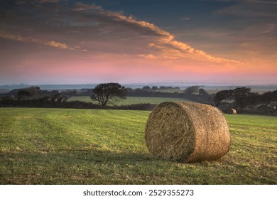 A beautiful, peaceful autumn scene in Cornwall, UK. The colours of sunrise fill the sky casting soft light on the landscape below. A hay bale is in the foreground with misty fields in the distance. - Powered by Shutterstock