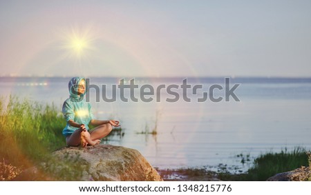 Beautiful peaceful woman sits in a pose of a half lotus practicing yoga meditation, blue summer sky, glowing chakra, eyes closed calm. Kundalini energy exercise. Human spirit, people healing concept