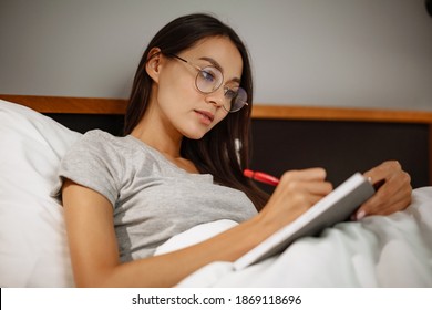Beautiful pensive asian woman writing notes while lying in bed