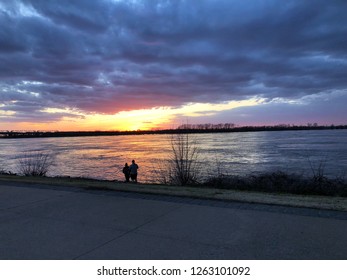 A Beautiful Photo Of A Sunset At Tom Lee Park In Memphis, TN. A Silhouetted Couple Stands Admiring The Sunset, On The Sand Beach Of The River.