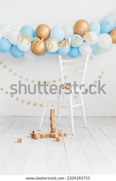 Beautiful photo zone for a birthday with blue and\
white balloons and the inscription Happy Birthday on a white wall.\
White chair with wooden eco toys. Wooden cubes and a wooden\
children\'s car