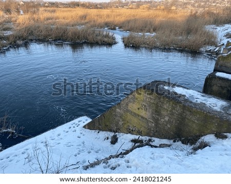 A beautiful picture of a river and a bridge in winter. The horizontal photo shows the shore, vegetation, reeds, water, snow, ice, structure, kami, footprints in the snow. A sunny cold day.