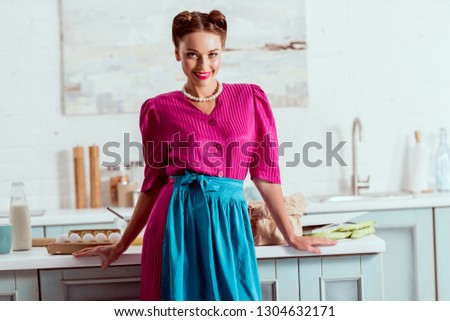 Beautiful pin up girl standing in light blue kitchen near table with different products and looking at camera